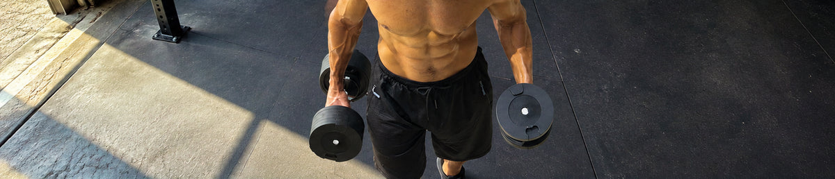 A man holding a snode dumbbell in each hand, standing in a gym on a dark concrete floor.